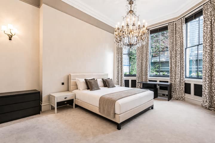 Një dhomë gjumi e qetë dhe luksoze me një krevat dopio "king", një llambadar të madh dhe dritare të larta me kanata të veshura me perde elegante, duke krijuar një vend të rafinuar plot dritë natyrale.