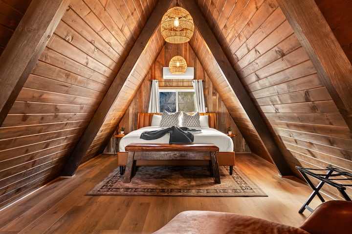 Another view of the loft bedroom showcasing the dramatic A-frame ceilings. King bed with Four Seasons–style bedding, nightstands with charging stations on both sides, and luggage racks for added comfort and convenience.
