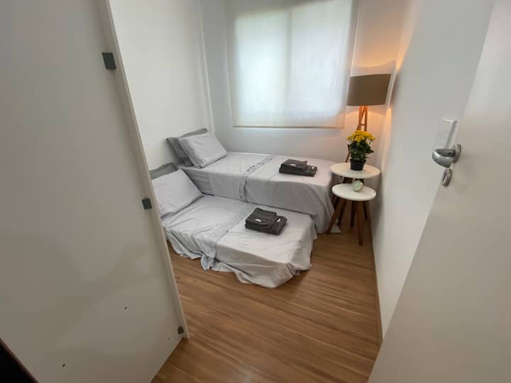 chambre simple avec deux lits, deux tables de chevet, un lampadaire, une horloge de table, une armoire, un ventilateur de plafond, un rideau à rouleau, une planche à repasser et un fer à repasser.