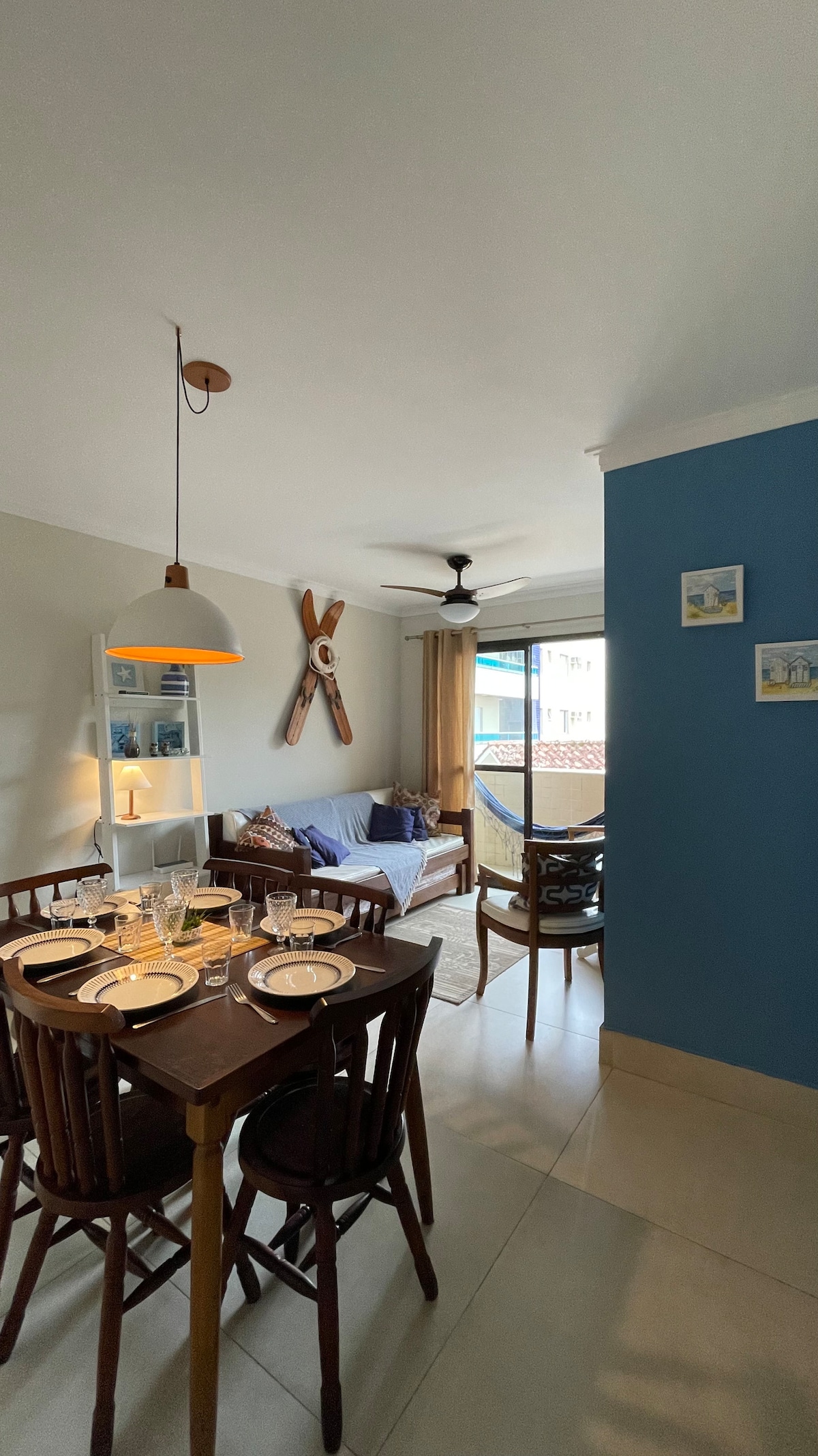 A dining area features a polished wooden table set for four, with plates and glasses arranged neatly. A cozy seating area is visible, highlighted by a plush sofa with decorative cushions. Natural light enters through a nearby window, enhancing the welcoming ambiance.