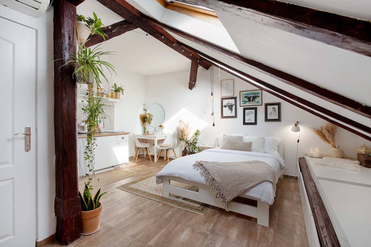 An open studio loft features a spacious bed with light linens, framed artwork on the walls, and a desk area with a round mirror. Plants are positioned in pots throughout the space, and exposed wooden beams add character to the ceiling.