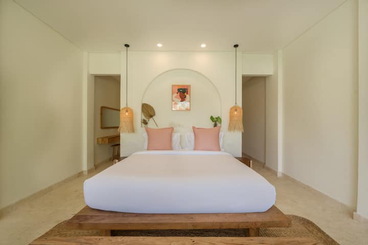Comfortable guest bedroom with crisp white bedding, soft pink pillows, and stylish hanging pendant lights. Open doors lead to a convenient walk-in closet with a dressing table and wall mirror.