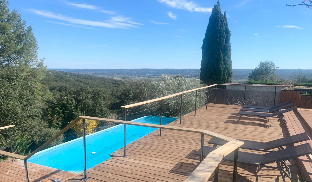 A wooden terrace overlooks the horizon, featuring a turquoise pool that blends with the landscape. Loungewear chairs are arranged nearby, providing space for relaxation. Lush greenery and trees contribute to the serene environment, enhancing the outdoor experience in a calm setting.
