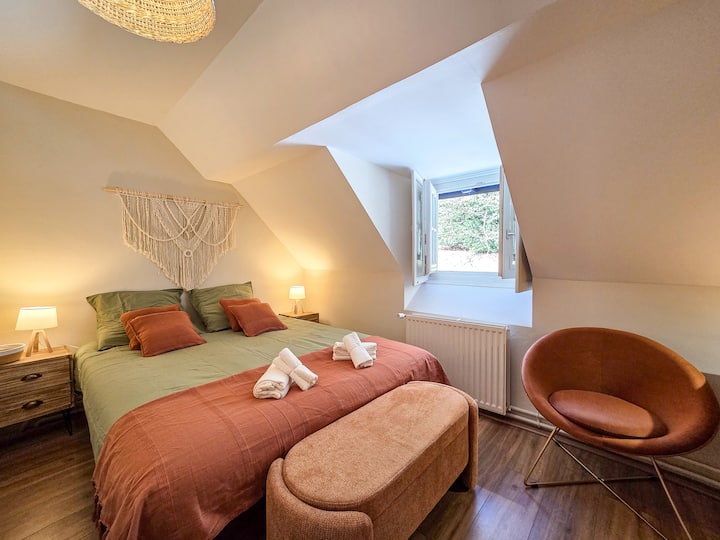 Habitación cálida con cama de 160 cm y mesitas de noche con lámparas. Delante, un baúl y un sillón añaden comodidad. La ventana con un radiador debajo y los tonos verdes y marrones aportan luz y ambiente cálido.