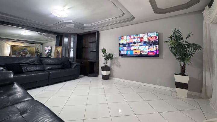 Sala elegante com sofá de couro preto, TV grande na parede com mosaico colorido, espelhos, prateleiras, lustre, piso branco e plantas em vasos. Moderna e aconchegante.