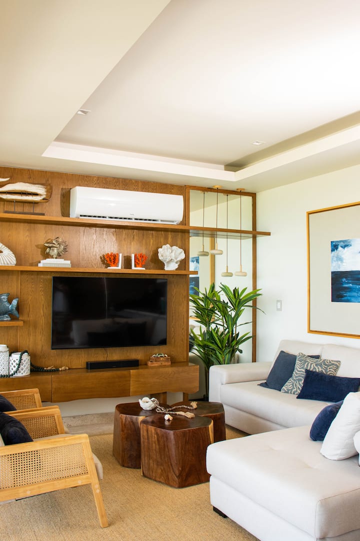 Sala de estar luminosa y acogedora, ideal para relajarse. Destaca por su elegante sofá blanco, sillones de mimbre con estilo tropical y mesas de centro de madera natural. Un espacio perfecto para descansar.
