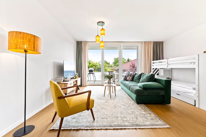 Wohnbereich mit Schlafsofa (180×200 cm), Sessel, Smart-TV, bodentiefen Fenstern und direktem Balkonzugang. Etagenbett in einer gemütlichen Ecke integriert.