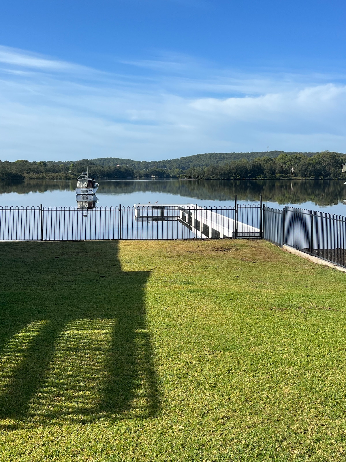 A serene water view is captured, showcasing a private jetty extending over calm waters. A fenced green lawn leads to the water's edge, with trees lining the opposite shore under a clear blue sky.
