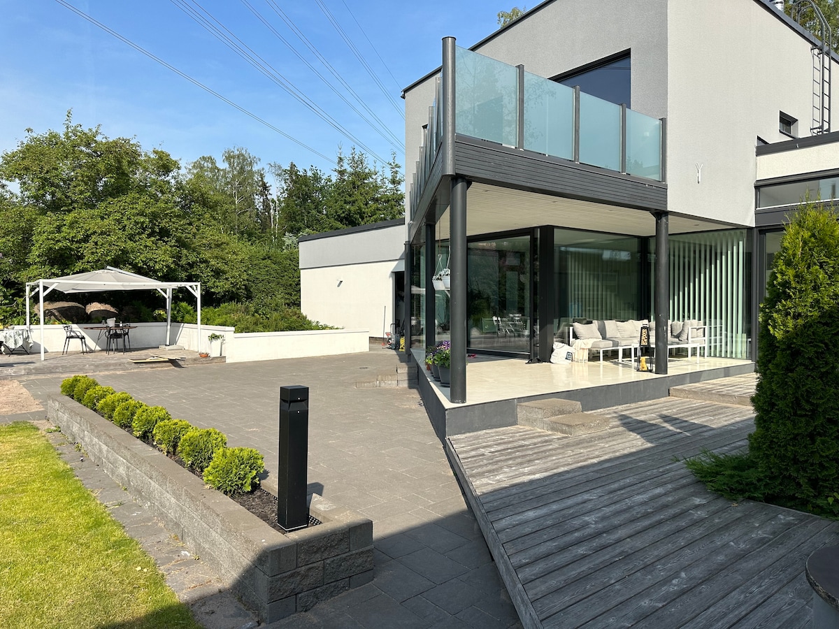 The exterior of the house features a modern design with large glass windows and a balcony. A spacious patio area is visible, equipped with seating options. Lush greenery surrounds the property, and a gazebo is present, providing additional outdoor relaxation space.