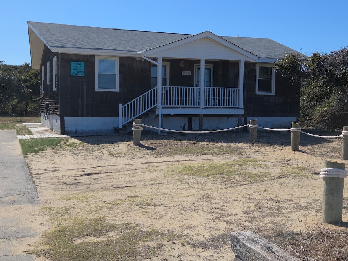 A beach cottage is depicted, featuring a front porch with white railings. The exterior is clad in dark wood, complemented by a gray roof. Sand surrounds the cottage, with a driveway visible on one side. Clear skies and bright sunlight illuminate the scene.