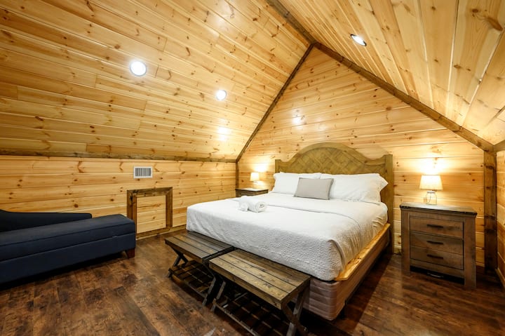 Este acogedor dormitorio en la cabaña con forma de A cuenta con techos abovedados de madera, una cómoda cama tamaño king y una iluminación ambiental cálida.