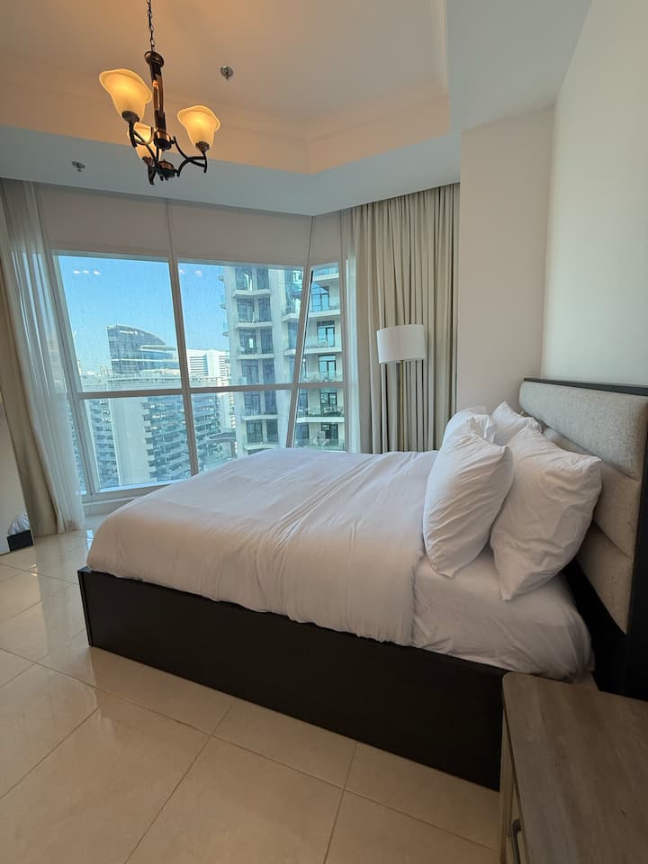 Habitación luminosa con suelos de baldosas brillantes, cama de tamaño completo con cabecero tapizado y sábanas, mesita de noche, lámpara de pie, lámpara de techo y grandes ventanas de esquina con cortinas que ofrecen vistas a la ciudad/balcón.