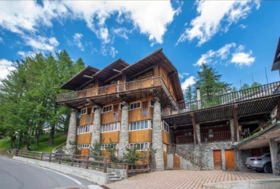 A charming multi-level building is surrounded by lush greenery, showcasing a blend of wood and stone in its architecture. The structure features balconies on upper levels and large windows, providing a warm, natural aesthetic. A paved driveway leads towards the entrance.