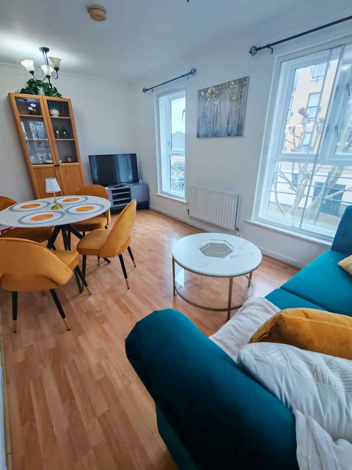 Világos nappali és étkező kékeszöld kanapéval, kényelmes párnákkal és egy négy személyes kerek étkezőasztallal. A nagy ablakok természetes fénnyel töltik meg a helyiséget, míg az ízléses dekoráció és a meleg hangulatú részletek barátságos légkört teremtenek a pihenéshez és a közös étkezésekhez.