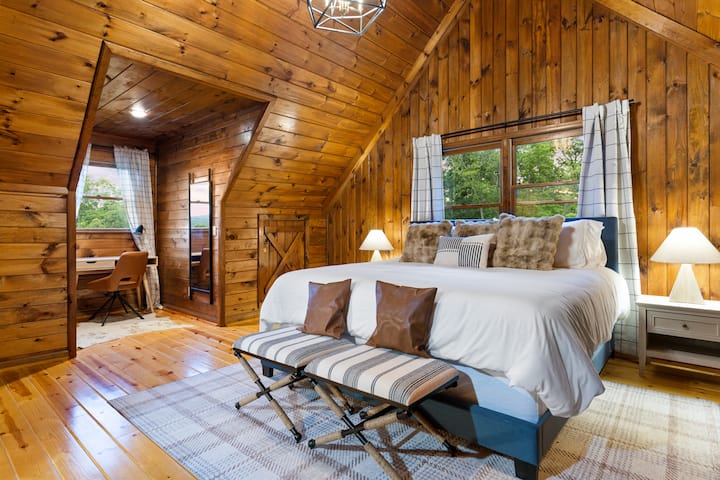 Relax in the cozy loft master bedroom featuring a plush king bed, warm wood interiors, and beautiful natural light from large windows. A dedicated workspace and peaceful cabin views make this the perfect retreat after a day of exploring