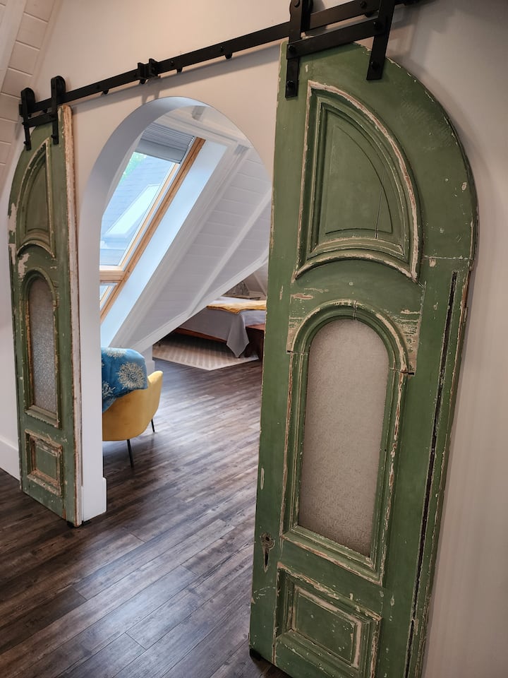 Puertas de granero arqueadas recicladas, únicas en su tipo, con pátina verde original y detalles vintage, que crean una entrada espectacular y memorable a la suite principal.