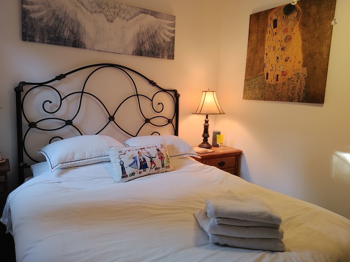 A cozy bedroom features a queen bed with white linens and decorative pillows. A bedside lamp emits a warm glow, complemented by an artwork on the wall. Fresh towels are neatly stacked on the bed, enhancing the inviting ambiance.