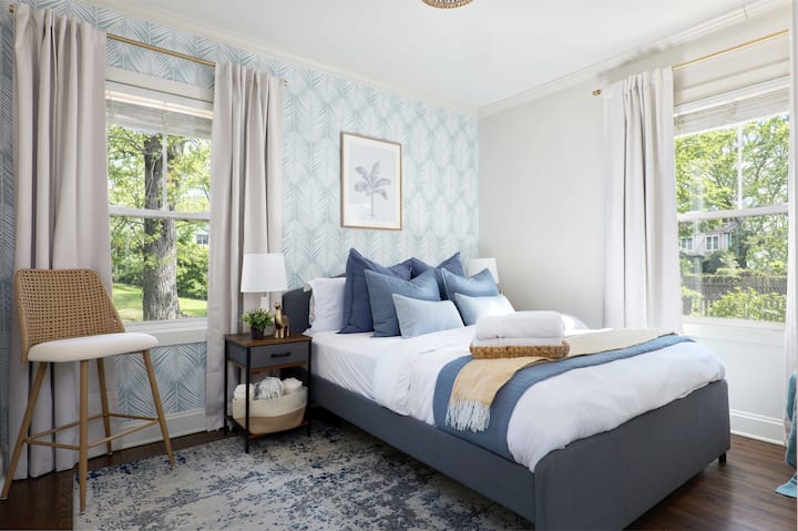 The second bedroom offers a bright, airy retreat with a plush queen bed and soft coastal tones. Large windows fill the space with natural light, creating a calm place to rest after a day in the city.