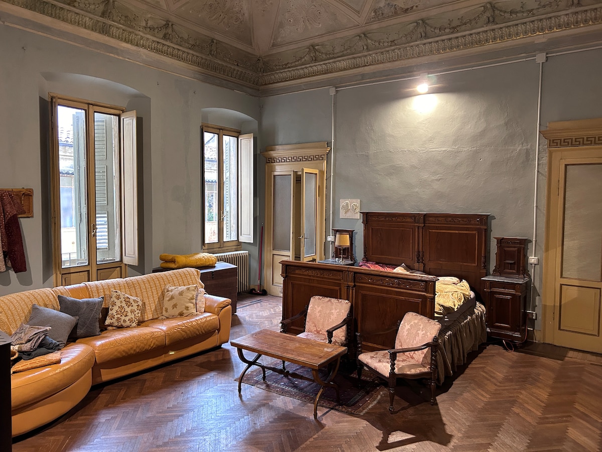 A spacious living area features large windows allowing natural light to fill the room. A curved orange sofa is complemented by a coffee table and wooden furniture, including a bed and dresser. Ornate details on the ceiling and wooden floors contribute to the historic charm.