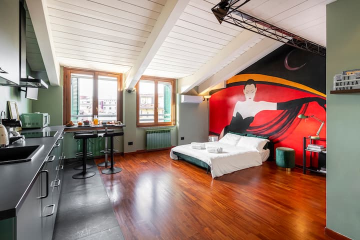Large open-plan space with an open kitchen, a dining area by the window, and a double sofa bed. Large artistic mural on the wall and warm wooden floors. Bright space with a sloping ceiling.