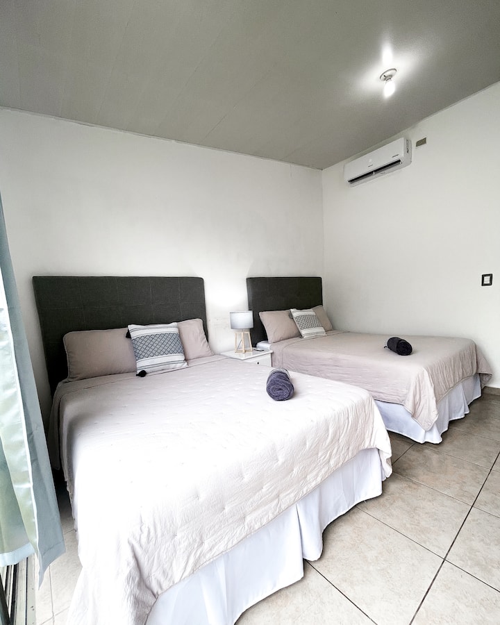 Habitación amplia y confortable con dos camas matrimoniales, sábanas de algodón, aire acondicionado y clóset. Cuenta con una amplia ventana y puerta corrediza con acceso al jardín, permitiendo la entrada de luz natural y aire fresco.