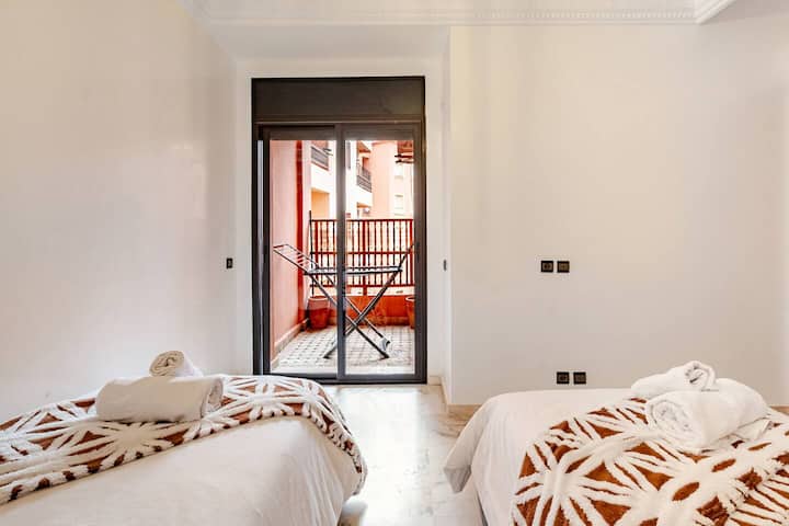 Chambre lumineuse et climatisée dotée de deux lits simples avec des couvre-lits et des oreillers à motifs orange, d'une grande armoire en bois et de portes coulissantes en verre qui s'ouvrent sur un balcon privé avec un séchoir à linge.