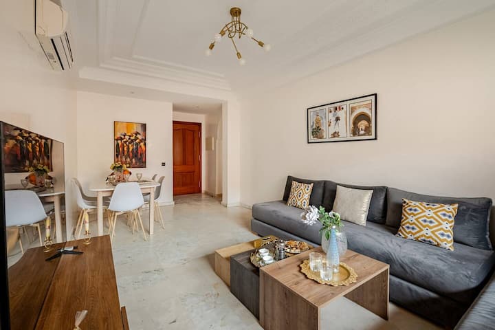 Salon climatisé avec un canapé gris en L, des coussins à motifs et une table basse en bois, à côté d'une fenêtre donnant sur un balcon.
