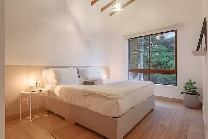 Habitación moderna y minimalista con techos altos y vigas de madera. Espacio lleno de luz natural, diseño sereno y cama premium para un descanso total. Ubicación tranquila, perfecta para recargar energías con estilo.