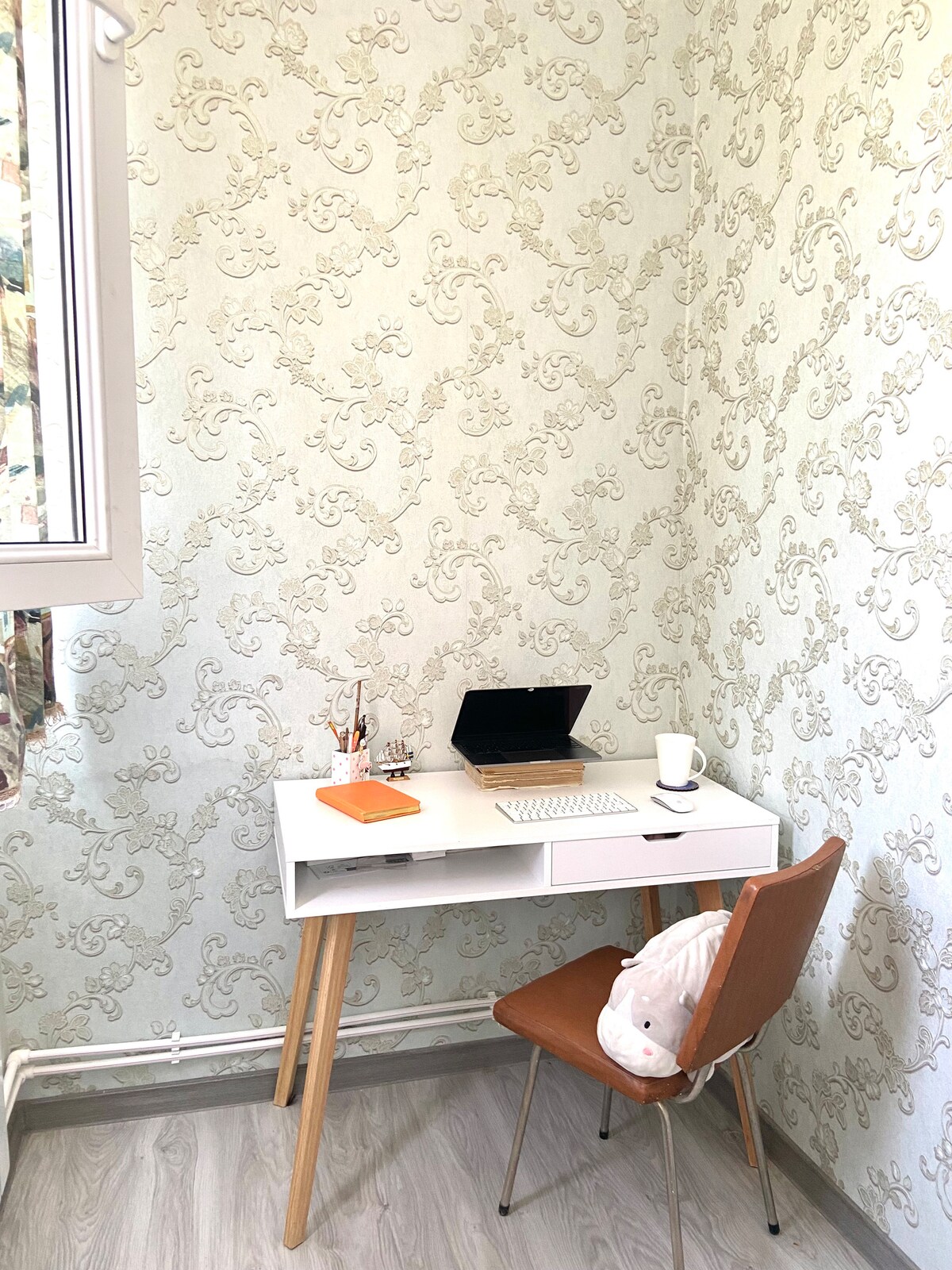 A compact workspace features a light wooden desk with two drawers, accompanied by a tan chair. A small laptop and a cup are positioned on the desk, while decorative stationery and a notebook enhance the setup. The wallpaper exhibits a delicate floral pattern.