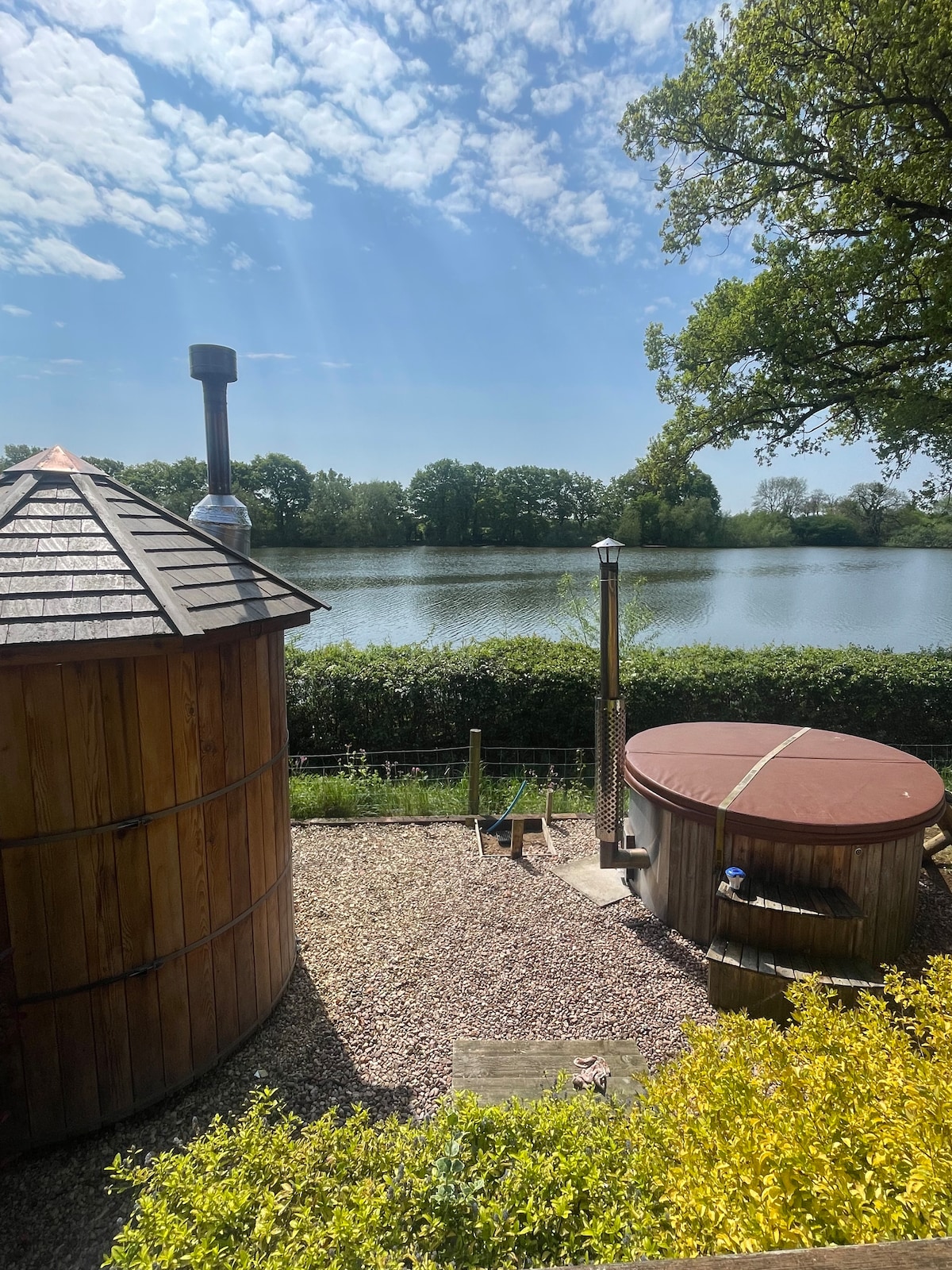 A tranquil scene showcases a hot tub beside a wooden structure, overlooking a serene reservoir. Lush greenery lines the water's edge, while a bright sky filled with soft clouds illuminates the area, creating a calming outdoor retreat.
