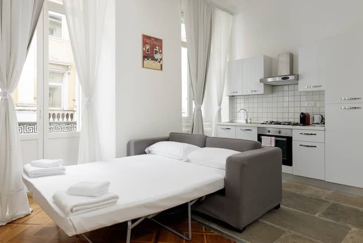 Sala con sofá cama, cocina y ventanas luminosas.