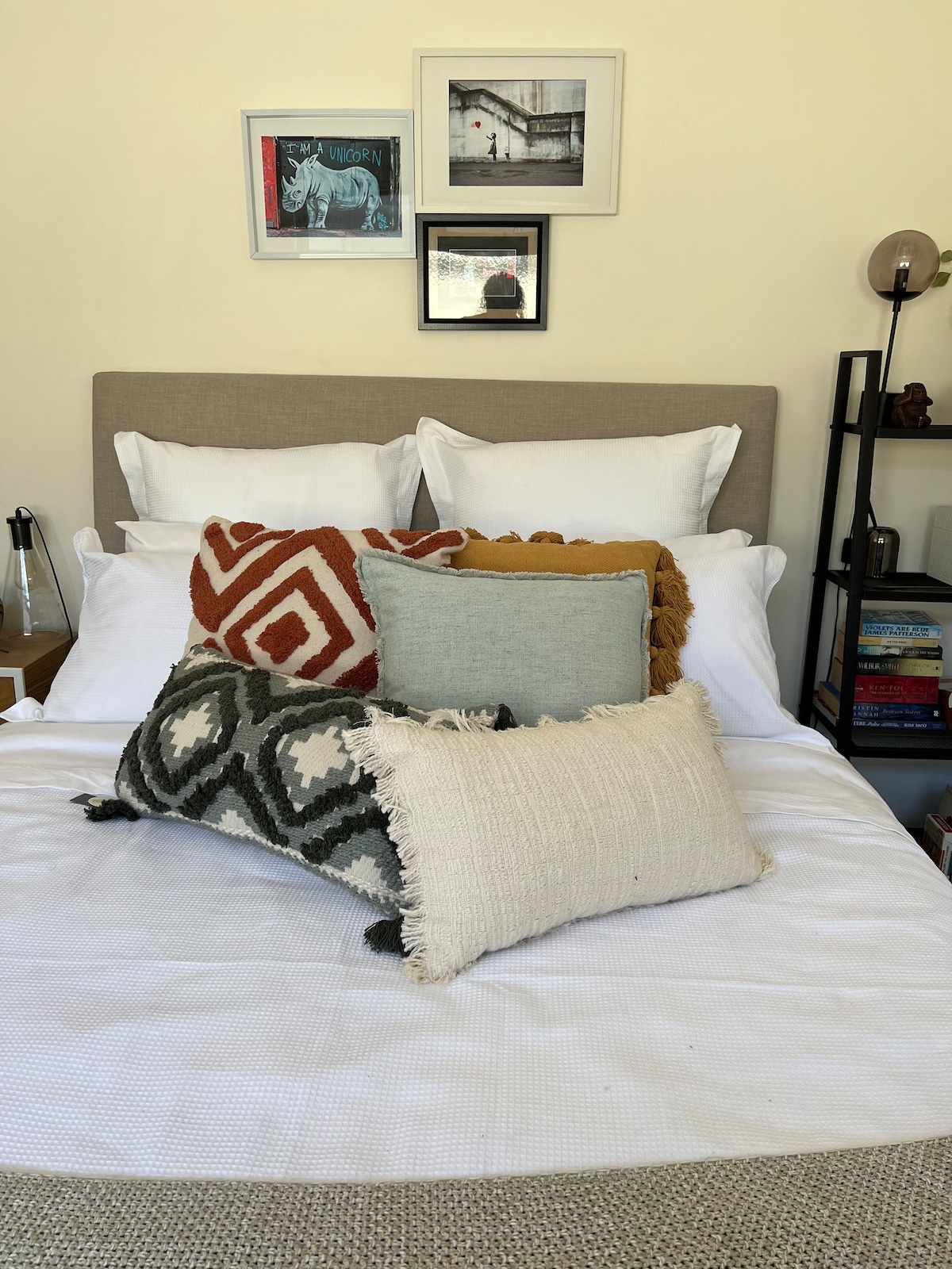A large, comfortable bed is adorned with several decorative pillows in various textures and colors, including beige, orange, and blue. The headboard is neutral, complemented by framed art and photographs positioned above the bed. A bookshelf is visible near the bedside.