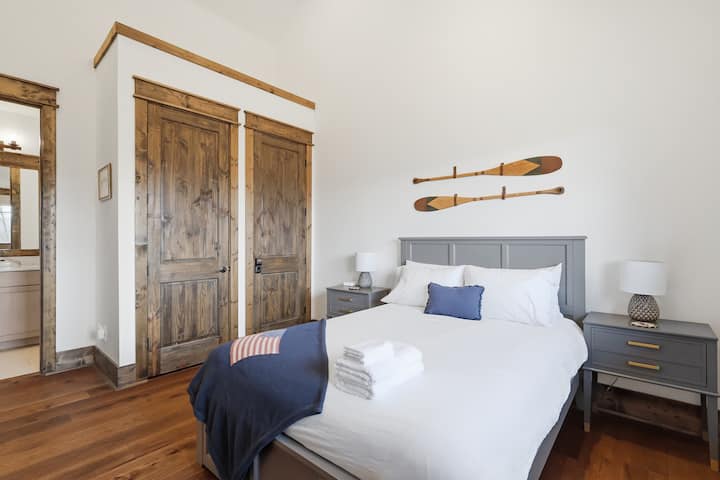 El dormitorio principal combina sábanas blancas inmaculadas con una divertida decoración de remos de madera y puertas corredizas de granero, lo que crea un ambiente acogedor que combina la montaña con la costa.