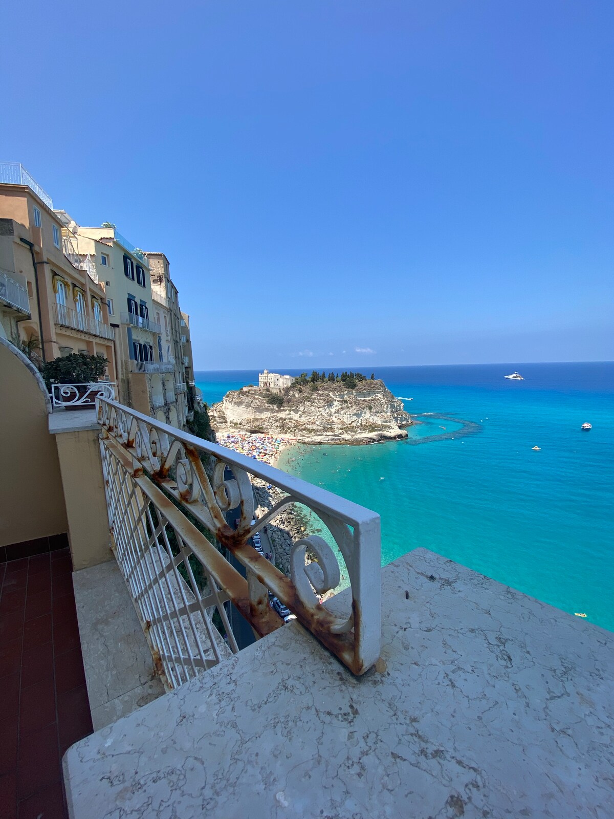 A spacious balcony offers stunning views of the sea, with bright turquoise waters and a small beach area visible below. Nearby, a scenic island features gentle greenery, while the coastline is lined with charming buildings. The clear blue sky enhances the tranquil atmosphere.