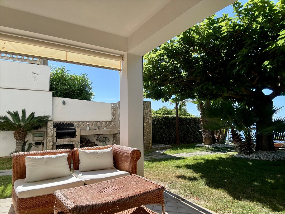 A shaded patio area features a woven seating arrangement and a low table. Lush greenery surrounds the space, with a barbecue grill visible in the background. Sunlight filters through the trees, enhancing the serene outdoor environment adjacent to the garden.