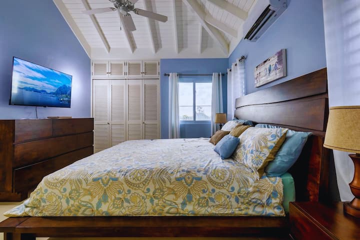 ห้องนอนที่สว่างพร้อมเพดานโค้งสูงและคานสีขาวที่มองเห็นได้ มีเตียงขนาดใหญ่พร้อมผ้าปูที่นอนลายสีน้ำเงินและสีเหลือง ตู้เสื้อผ้าและหัวเตียงไม้สีเข้ม และทีวีติดผนัง หน้าต่างที่ให้แสงธรรมชาติและวิวของต้นไม้เขียวชอุ่ม