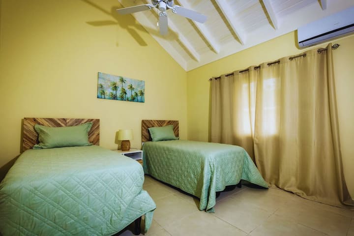 ห้องพักน่าอยู่ที่มีแสงสว่างดี พร้อมผนังสีเหลืองอ่อนและเพดานโค้งสูง มีเตียงแฝด 2 เตียงที่สะดวกสบายพร้อมผ้าปูที่นอนสีเขียวเซจ หัวเตียงไม้สีเข้ม และโต๊ะข้างเตียงตรงกลาง ม่านผืนใหญ่และพัดลมเพดานช่วยให้มั่นใจได้ว่าจะได้บรรยากาศที่เย็นสบายและผ่อนคลาย