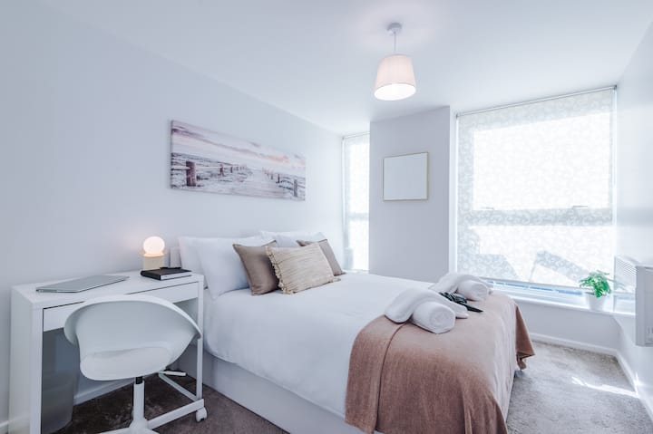 Светла и минималистична спалня с бяло двойно легло с възглавници в неутрални тонове, изчистено бяло бюро с лаптоп и големи прозорци, които изпълват помещението с естествена светлина.