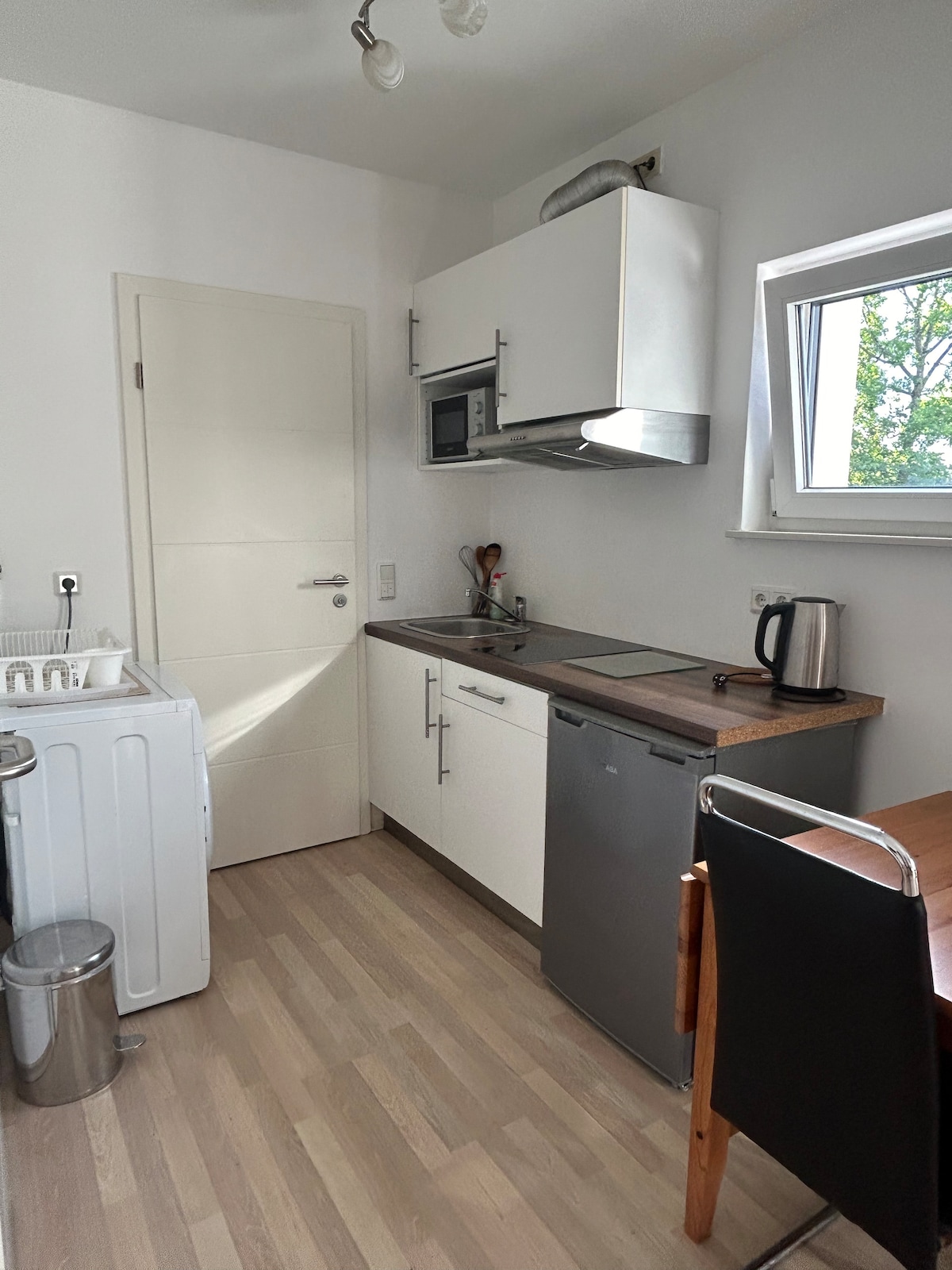 The kitchen area is equipped with modern appliances, including a microwave, kettle, and dishwasher. A washing machine is positioned beside the countertop. Natural light enters through a window, illuminating the clean white cabinetry and warm wooden tones of the floor.