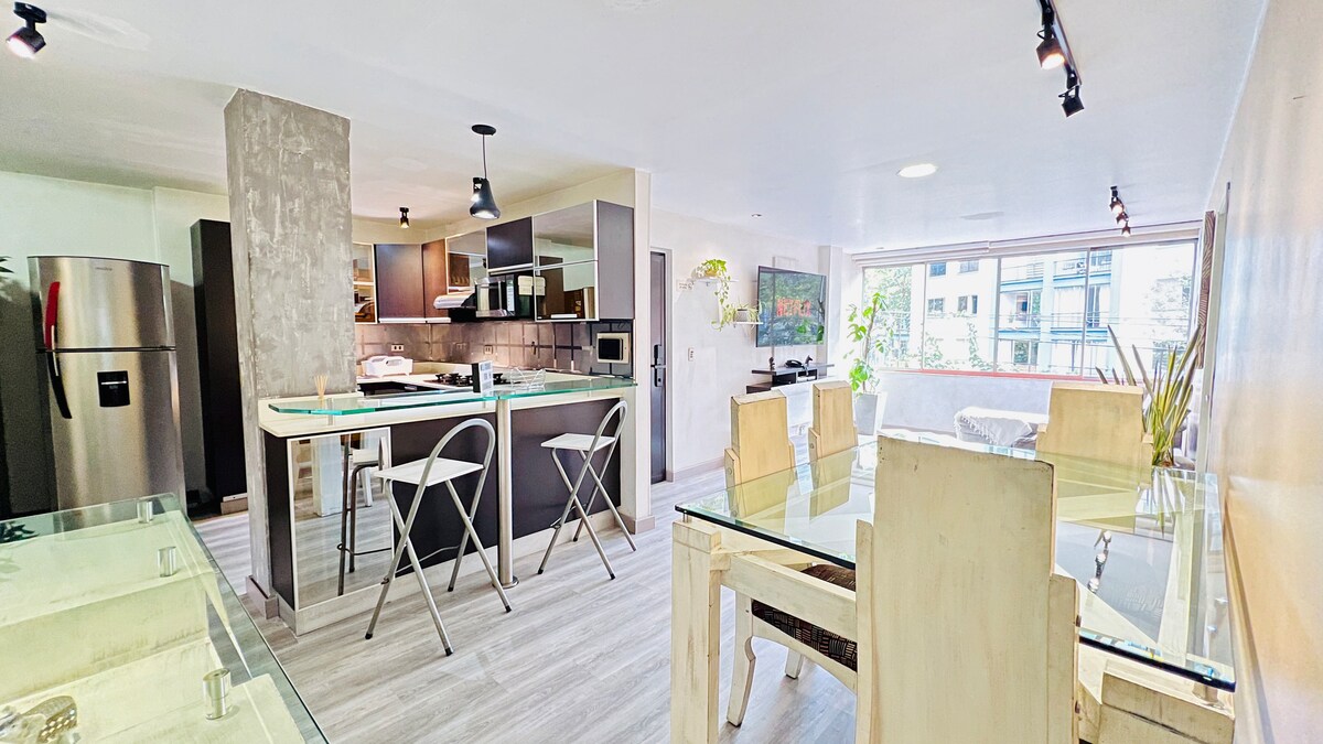 An open-plan living area showcases a modern kitchen with wooden cabinetry and stainless steel appliances. A glass dining table is set for six, surrounded by light-colored chairs. Natural light fills the space through a large window, enhancing the airy atmosphere.