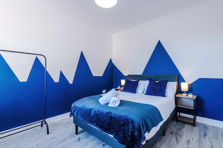 Dormitorio luminoso y moderno con una cómoda cama doble, iluminación junto a la cama y paredes en un llamativo azul para una estancia con estilo.