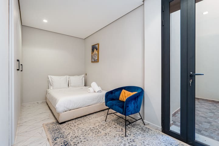 Une chambre lumineuse et moderne comprend un lit blanc avec des draps frais, un fauteuil en velours bleu et une porte vitrée à cadre foncé donnant sur un espace extérieur.