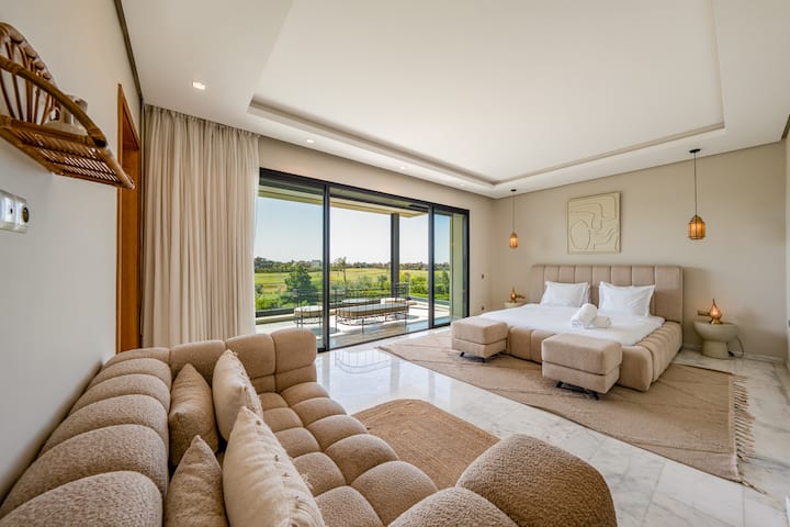 Une chambre moderne et lumineuse est dotée d’un grand lit, d’un canapé incurvé et d’un balcon offrant une vue sur le terrain de golf. La pièce a un décor clair et des planchers en marbre. Ajouter une description visuelle.