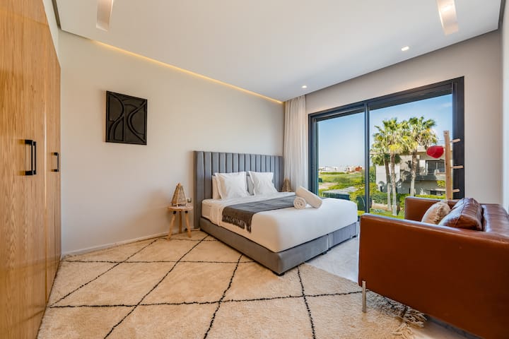 Une chambre lumineuse et moderne avec un grand lit, un fauteuil brun et un tapis à motifs. Une grande fenêtre donne sur des palmiers et offre beaucoup de lumière naturelle.