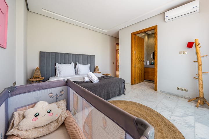 Une chambre lumineuse et moderne dotée d’un grand lit avec des draps blancs, d’un portemanteau en bois et d’un lit pour bébé au premier plan. Une porte mène à une salle de bain attenante.