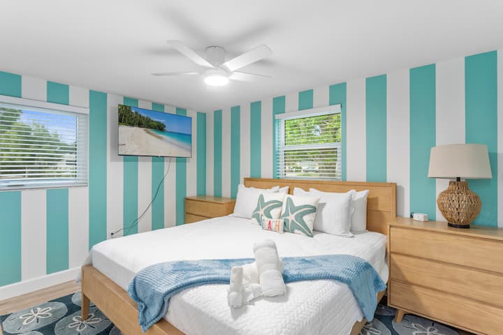 Dormitorio de inspiración costera con una cómoda cama king, decoración de estilo playero y detalles en tonos azules frescos. Disfrute de un refugio relajante con televisión, luz natural y detalles limpios y modernos para una estancia tranquila.