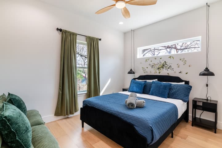 Cette chambre paisible est dotée d'un lit douillet avec des draps bleu foncé et d'un mur à motif floral saisissant, le tout encadré par de grandes fenêtres aux élégants rideaux vert olive.
