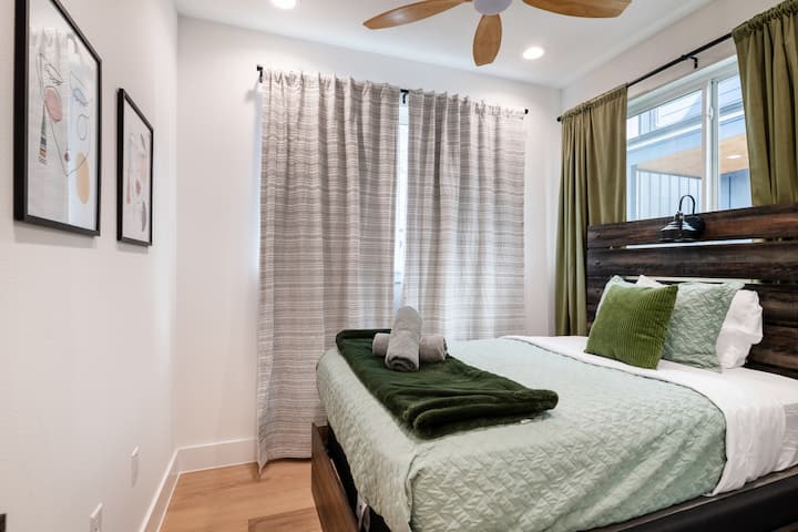 Cette chambre paisible est dotée d'un lit queen confortable avec des touches de vert douces, d'un ventilateur de plafond moderne en bois et de grandes fenêtres avec des rideaux d'intimité pour un séjour reposant.