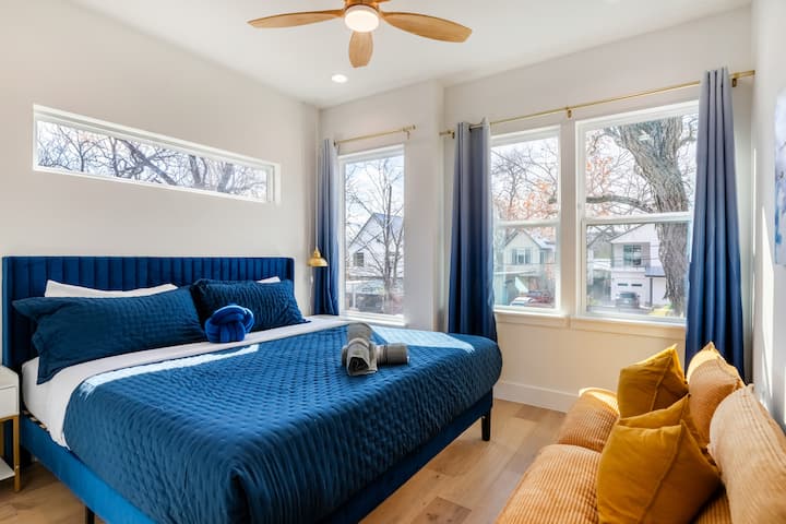 Cette chambre accueillante est dotée d'un grand lit très confortable avec des draps bleu roi et d'un canapé jaune vif, le tout baigné de lumière naturelle grâce à plusieurs grandes fenêtres.
