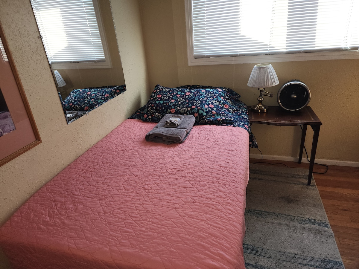 A private bedroom is presented with a twin bed, covered in a light pink quilt. Colorful floral-patterned pillows rest against the wall. A small nightstand holds a lamp and a fan, with natural light streaming through partially opened blinds.